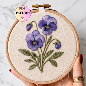 Puede incluir: Un bastidor de bordado con un diseño floral detallado de pensamientos morados con centros amarillos y hojas verdes. El bordado está sobre una tela beige, y el bastidor es de madera. El texto "PDF PATTERN" es visible.