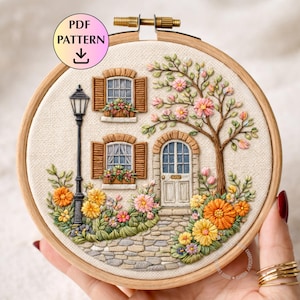 Puede incluir: Arte de aro bordado que presenta una escena detallada de una casa de campo. El diseño incluye una casa con contraventanas, una puerta y un árbol con flores rosas. Un camino de piedra y flores coloridas rodean la casa. El texto "PDF PATTERN" es visible.