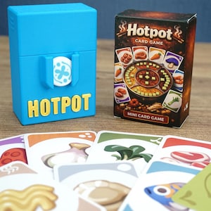 Könnte beinhalten: Eine hellblaue Kunststoff-Aufbewahrungsbox mit dem Wort HOTPOT in gelben Buchstaben neben einer Hotpot-Kartenspielschachtel mit einer Grafik eines Hotpots und verschiedenen Zutaten sowie mehreren ausgebreiteten Spielkarten.