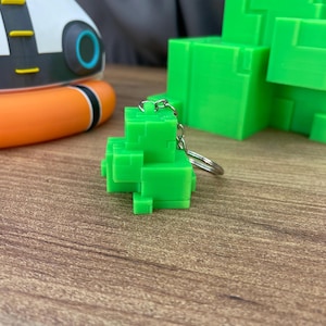 Op de afbeelding: Een felgroene sleutelhanger met een blokvormige, gepixelde structuur bestaande uit meerdere kleine kubussen. De sleutelhanger is bevestigd aan een zilverkleurige metalen ring en ketting en heeft een geometrisch ontwerp met scherpe randen.