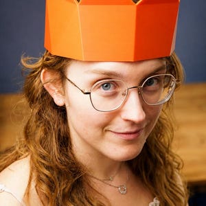 May include: A bright orange, geometric crown with a multi-pointed, angular design. The crown features a solid, smooth surface and a structured, rigid form that sits atop the head, showcasing a vibrant, uniform colour throughout the entire piece.