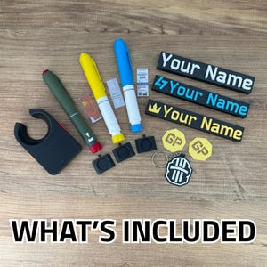 Puede incluir: Una colección de artículos que incluye un clip circular negro, tres bolígrafos en verde oliva, amarillo y azul, tres placas rectangulares con el texto Your Name, dos fichas hexagonales GP, tres cuadrados negros pequeños y un llavero en blanco y negro.