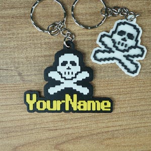 Può includere: Due portachiavi con disegni di teschio e ossa incrociate. Un portachiavi ha uno sfondo nero con la scritta "Your Name" in giallo. L'altro ha un disegno di teschio e ossa incrociate bianche con un contorno nero.