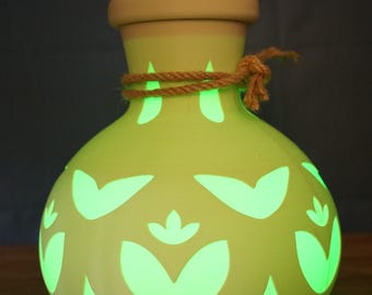 May include: A light yellow, rounded ceramic lamp with a smooth finish and a thick rim. The body features cut-out floral patterns that emit a bright green glow from within. A piece of natural brown twine is tied around the neck of the lamp.