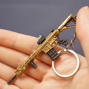 Gold rifle keyring held in hand with key loop attached, showing detailed miniature gaming charm from the side.