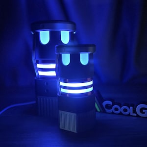 Puede incluir: Dos dispositivos electrónicos cilíndricos con cuerpos de color gris oscuro y acentos de luz azul brillante. Las luces forman dos rectángulos verticales redondeados en la parte superior y dos bandas horizontales en el centro. El texto COOLG es visible.