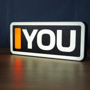 May include: A rectangular LED sign with a black textured frame and a white border. The sign features a vertical orange rectangle on the left side followed by the white text YOU in a bold sans-serif font against a black background.