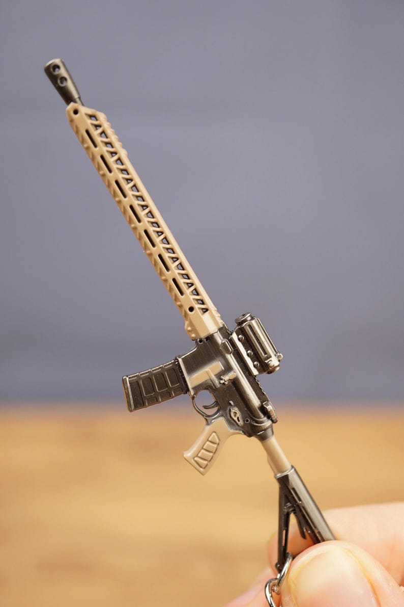 Close-up of tan and black tactical rifle keyring held by fingers, showing long barrel, sight and magazine details.