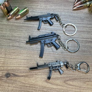 May include: Three miniature black metal submachine gun keychains with attached silver-toned split rings. Each keychain features a detailed replica of a firearm with a magazine, trigger, and barrel. Several brass-colored bullet casings are scattered nearby.