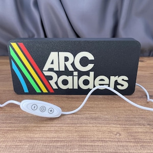 May include: A rectangular black LED sign featuring the white text ARC Raiders and a diagonal rainbow stripe pattern on the left side. The sign includes a white power cord with an integrated control panel containing power and brightness buttons.