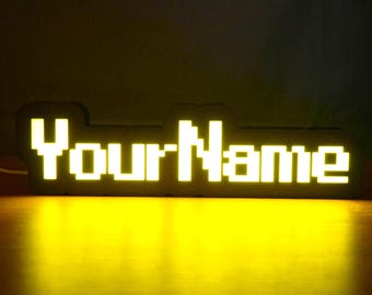 May include: A custom LED sign featuring the text YourName in a blocky, pixelated font. The letters are illuminated with a bright yellow light, creating a glowing effect against the dark, solid background of the sign's frame structure.