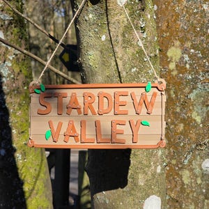 Könnte beinhalten: Ein rechteckiges beiges Schild mit dem Text STARDEW VALLEY in erhabenen braunen Buchstaben. Das Schild hat kleine grüne Blattakzente an den Ecken und ist an einer dünnen Schnur aufgehängt, mit einem horizontalen Planken-Design und sichtbaren Nieten.