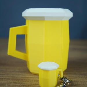 Könnte beinhalten: Ein gelb-weißer, geometrischer Bierkrug mit einem passenden Miniatur-Bierkrug-Schlüsselanhänger. Der größere Krug hat ein facettiertes Design mit weißem Rand und stabilem Henkel, während die kleinere Schlüsselanhänger-Version einen silbernen Karabiner hat.