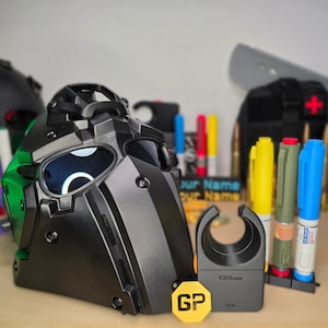 May include: A black tactical helmet with a dark visor, positioned next to a black CGS-branded device, a yellow hexagonal GP token, and three upright markers in yellow, olive green, and blue. The background features blurred office supplies.