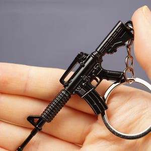 Black tactical rifle keychain close-up with metal key loop, designed as a compact FPS gamer backpack charm.