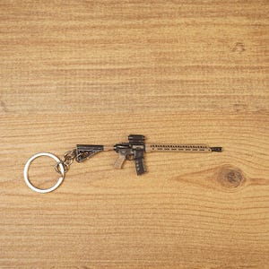 Tan and black long rifle keychain with metal key loop displayed on a wooden tabletop for tactical shooter fans.