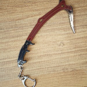 Puede incluir: Un llavero en miniatura con forma de piolet que presenta un mango de color rojo oscuro y una cabeza de metal plateado con una punta dentada y una pala. El agarre es negro y texturizado, unido a una anilla metálica plateada por una cadena.