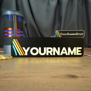 Könnte beinhalten: Ein rechteckiges schwarzes Schild mit strukturierter Oberfläche, das den Text YOURNAME in weißen Großbuchstaben und ein mehrfarbiges Regenbogenstreifen-Motiv auf der linken Seite zeigt. Ein kleineres passendes Schild zeigt COOLGAMINGSTUFF.