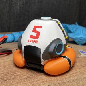May include: A white, gray, and black spherical lifepod model featuring the red number 5 and the text LIFEPOD printed on the front. The object includes bright orange inflatable-style side components and small yellow accents along the top edge.