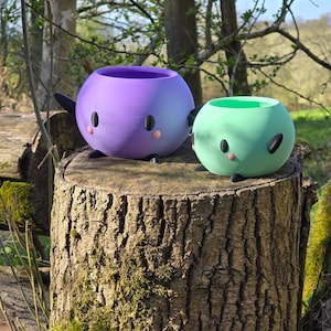 May include: Two round planters with smooth surfaces, one larger purple and one smaller mint green. Each features two black oval eyes, a small pink circular nose, and two small black feet extending from the base, creating a character-like appearance.