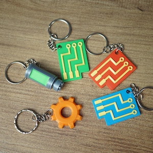 Pode incluir: Uma coleção de chaveiros com vários designs. Inclui um cilindro verde, uma engrenagem laranja e três chaveiros retangulares em verde, vermelho e azul, cada um com padrões de circuitos amarelos. Todos os chaveiros têm argolas prateadas.