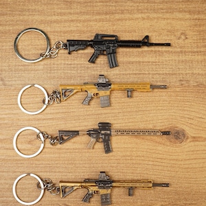 Four miniature tactical rifle keyrings in black, gold and tan styles laid flat on a wooden surface for gamer gift display.