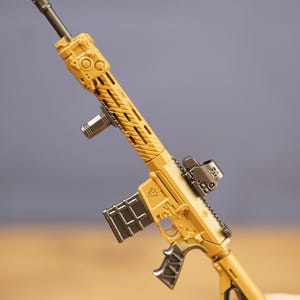 Close-up of tan tactical rifle keychain held between fingers showing detailed barrel, sight, magazine and grip.