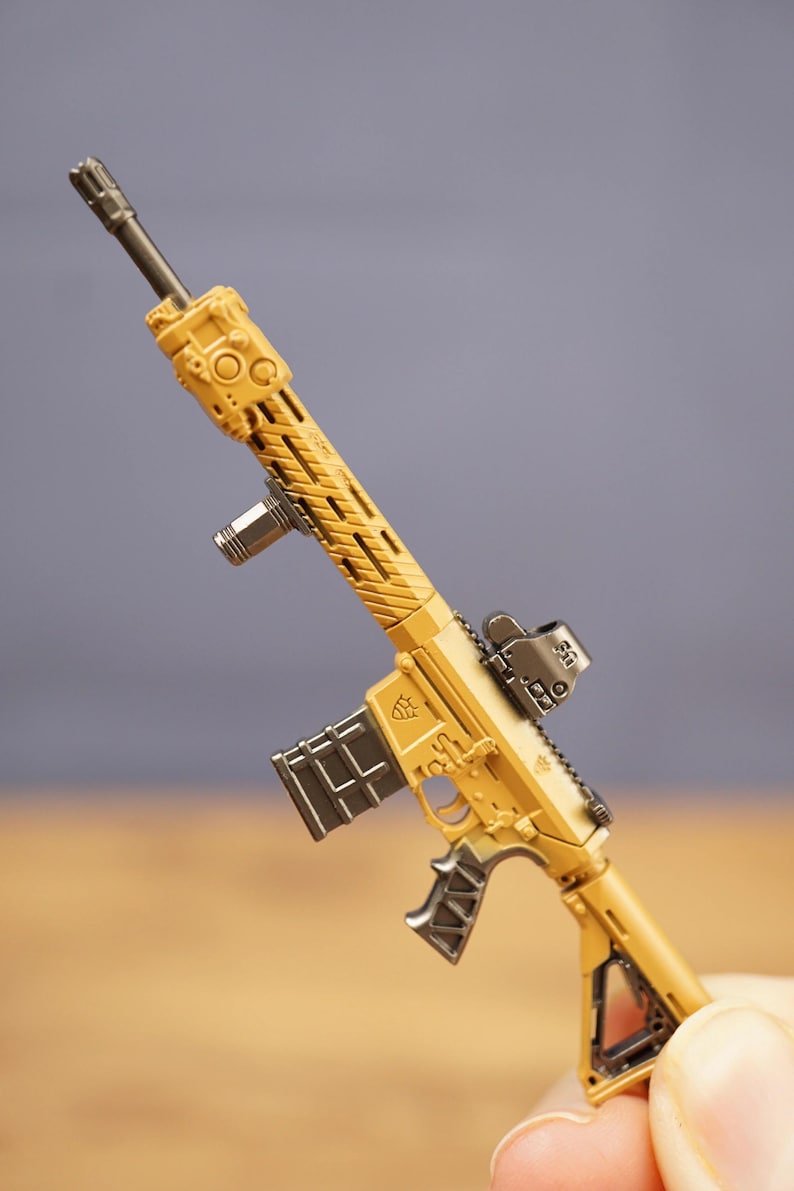 Close-up of tan tactical rifle keychain held between fingers showing detailed barrel, sight, magazine and grip.