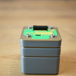 Könnte beinhalten: Eine quadratische, mehrschichtige elektronische Komponente mit einer grauen Basis, einem goldfarbenen Mittelteil und einer grünen Leiterplatte als Oberseite. Die Oberseite enthält einen schwarzen Anschluss und vier silberne Metallpunkte.