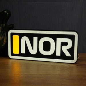 Puede incluir: Un letrero luminoso rectangular con fondo negro y borde blanco. El texto "INOR" aparece en letras mayúsculas blancas grandes, precedido por un rectángulo amarillo vertical en el lado izquierdo de la cara del letrero.