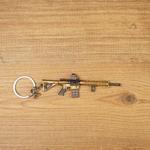 Gold tactical rifle keyring with detailed sight, magazine and barrel shown on wood as a gaming backpack charm.
