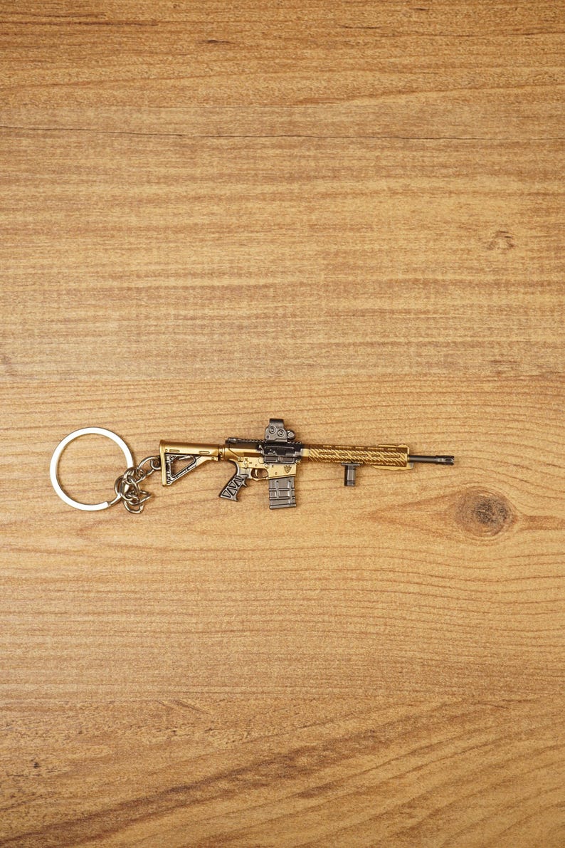 Gold tactical rifle keyring with detailed sight, magazine and barrel shown on wood as a gaming backpack charm.