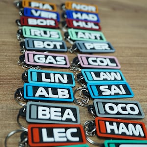 Peut inclure: Une collection de porte-clés rectangulaires en silicone de différentes couleurs, chacun comportant un texte blanc de trois lettres et un petit carré coloré sur la gauche. Les porte-clés sont fixés à des anneaux en métal argenté et disposés en deux colonnes.