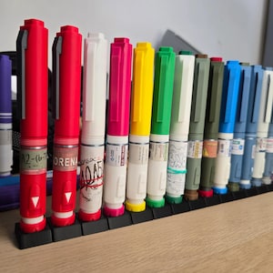 Puede incluir: Una colección de plumas autoinyectoras cilíndricas multicolores dispuestas en fila sobre un soporte negro. Las plumas presentan varios colores incluyendo rojo, blanco, rosa, amarillo, verde, azul y oliva, con etiquetas y texto impreso.