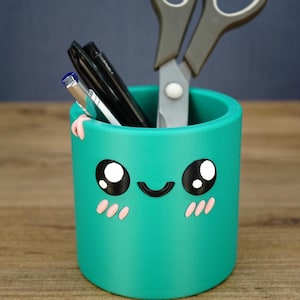 May include: A cylindrical teal desk organizer featuring a printed face with large black eyes, a curved smile, and pink blush marks. The container holds a pair of grey and black scissors along with several black and silver pens and markers.