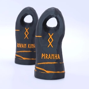 May include: Two dark grey, faceted, vertical objects with a hole at the top. Each features an orange geometric symbol and horizontal orange lines. One object is labelled with the text RIVAN KING and the other with the text PIRANHA in orange.