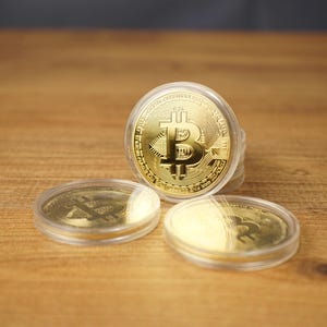 May include: Three gold-colored Bitcoin tokens encased in clear circular plastic protective capsules. The central token stands upright, displaying the Bitcoin symbol and the text "Bitcoin Digital Decentralized Peer to Peer" embossed on its surface.