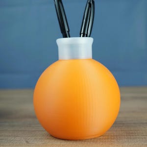 Könnte beinhalten: Ein orangefarbener Stifthalter mit kugelförmigem Boden und weißem Aufsatz, der zwei schwarze Stifte enthält. Der Stifthalter besteht aus einem strukturierten Material und steht auf einer Holzoberfläche. Der Hintergrund ist einfarbig blau.