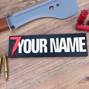 Pode incluir: Um emblema retangular preto com um número 7 vermelho estilizado e o texto branco YOUR NAME em letras maiúsculas em negrito. O emblema é exibido ao lado de uma lâmina serrilhada cinza, dois cartuchos de espingarda vermelhos e dois projéteis.