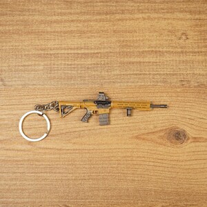 Tan and black tactical rifle keyring with detailed grip, sight and front attachment on a wooden background.