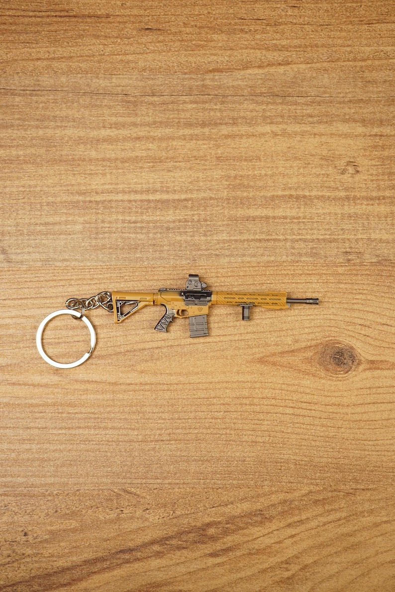 Tan and black tactical rifle keyring with detailed grip, sight and front attachment on a wooden background.