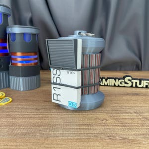 Op de afbeelding: Een cilindervormig grijs rekwisiet met een wit rechthoekig paneel met de tekst R1SS, R165 en CGS in het blauw. Het object heeft een geribbelde textuur, een metalen ring aan de bovenkant en wordt vastgehouden door twee donkere banden. Een tweede is zichtbaar.