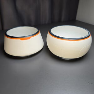 Puede incluir: Dos cuencos de color blanco roto con un acabado mate y liso. Cada cuenco presenta un diseño de borde distintivo que consiste en una fina banda negra sobre una franja naranja vibrante. Los cuencos tienen una forma redondeada y cónica.