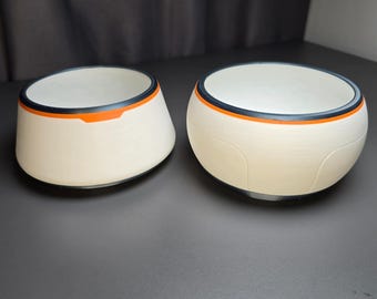 May include: Two off-white bowls with a smooth, matte finish. Each bowl features a distinct rim design consisting of a thin black band layered above a vibrant orange stripe. The bowls have a rounded, slightly tapered shape with a clean, simple aesthetic.