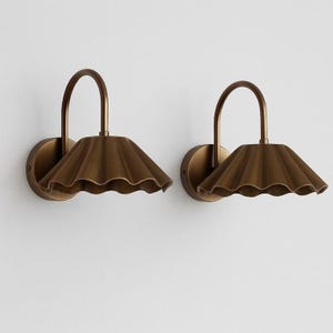 May include: A pair of brass wall sconces. Each sconce has a curved arm and a ruffled, umbrella-shaped shade. The sconces are mounted on circular backplates. The design is vintage-inspired. The sconces would suit a bedroom or living room.