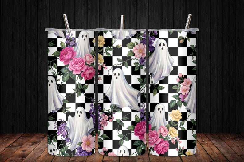 May include: A cylindrical tumbler with a straw featuring a repeating pattern of white ghosts, pink and yellow flowers, green leaves, and a black and white checkered background. The design wraps around the entire surface of the tall beverage container.