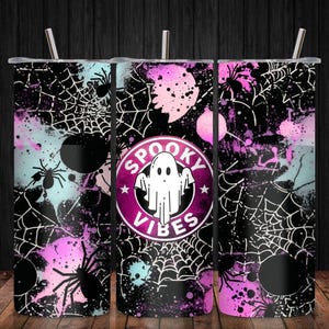 May include: A cylindrical stainless steel tumbler with a straw, featuring a black, pink, and light blue splatter pattern with spider web and spider graphics. The center displays a circular logo with a white ghost and the text SPOOKY VIBES.