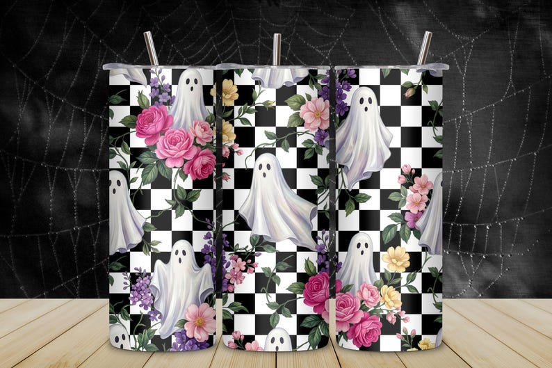 May include: A cylindrical tumbler with a straw featuring a repeating pattern of white ghosts, pink and yellow flowers, green leaves, and a black and white checkerboard background. The design covers the entire surface of the metal drink container.