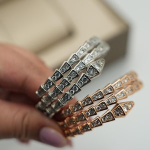 Puede incluir: Dos pulseras enrolladas con un diseño similar a una serpiente, una en metal plateado y otra en metal color oro rosa. Ambas pulseras están incrustadas con numerosas piedras pequeñas, claras y brillantes dispuestas en un patrón geométrico.
