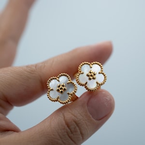 May include: A pair of gold-toned stud earrings shaped like five-petaled flowers. Each earring features a white mother-of-pearl inlay within a beaded gold-toned border, with a small cluster of gold-toned beads at the center of the floral design.